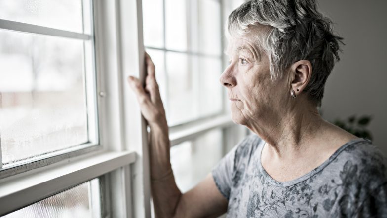An elderly white woman stands looking out of a window with her hand on the glass pane.